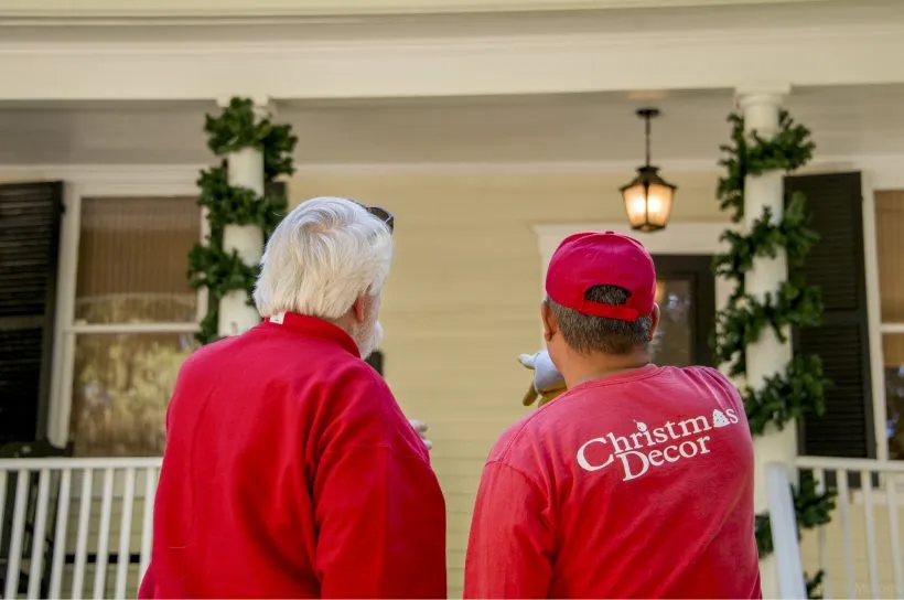 Two decorators in Christmas Decor red looking over a garlanded porch