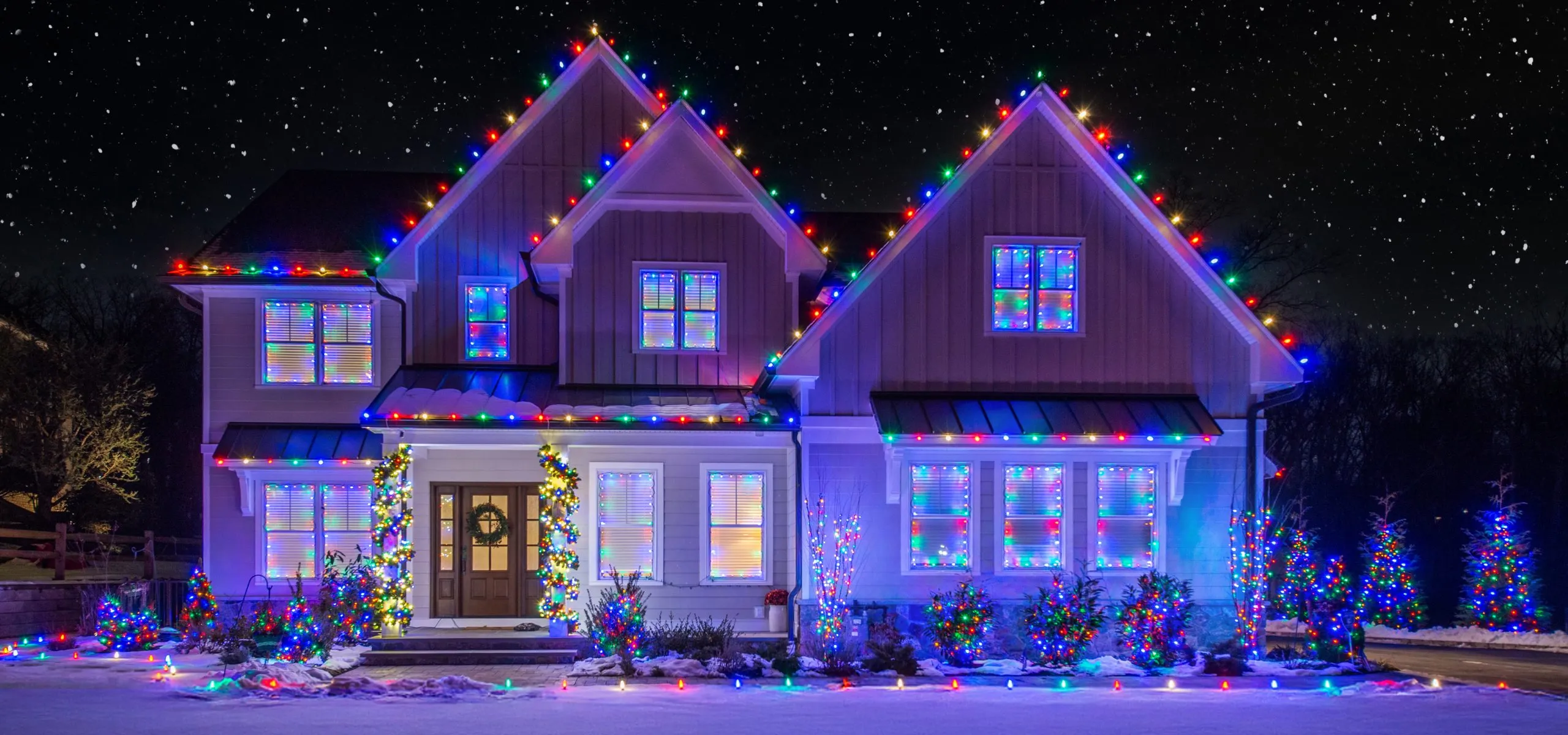 A two-story home lit in blue and purple, with matching lit trees along the drive