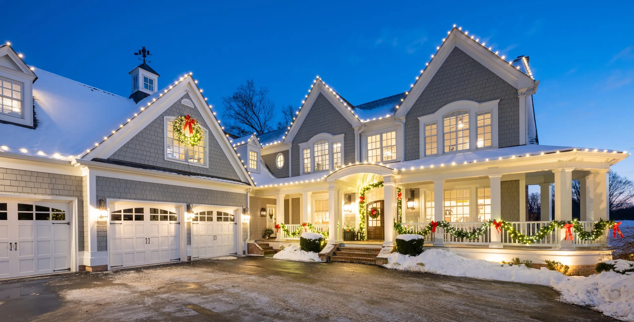 A shingled two-story home outlined in warm white lights at dusk, with lit wreaths and garland across the porch