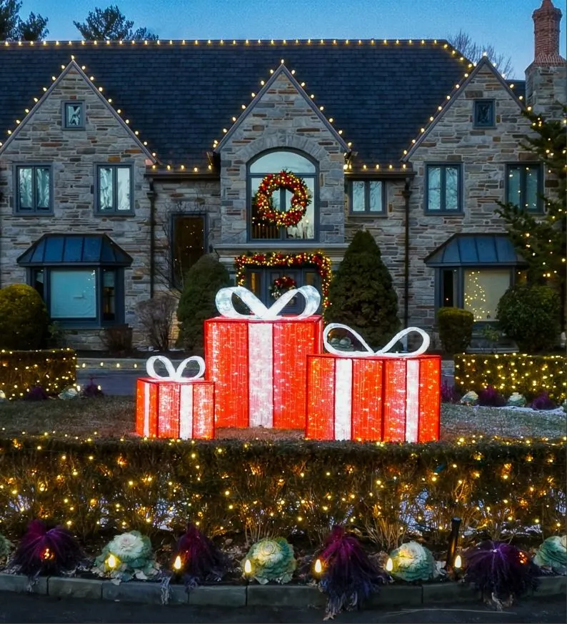 Lit gift boxes on the lawn of a stone house strung with roofline lights