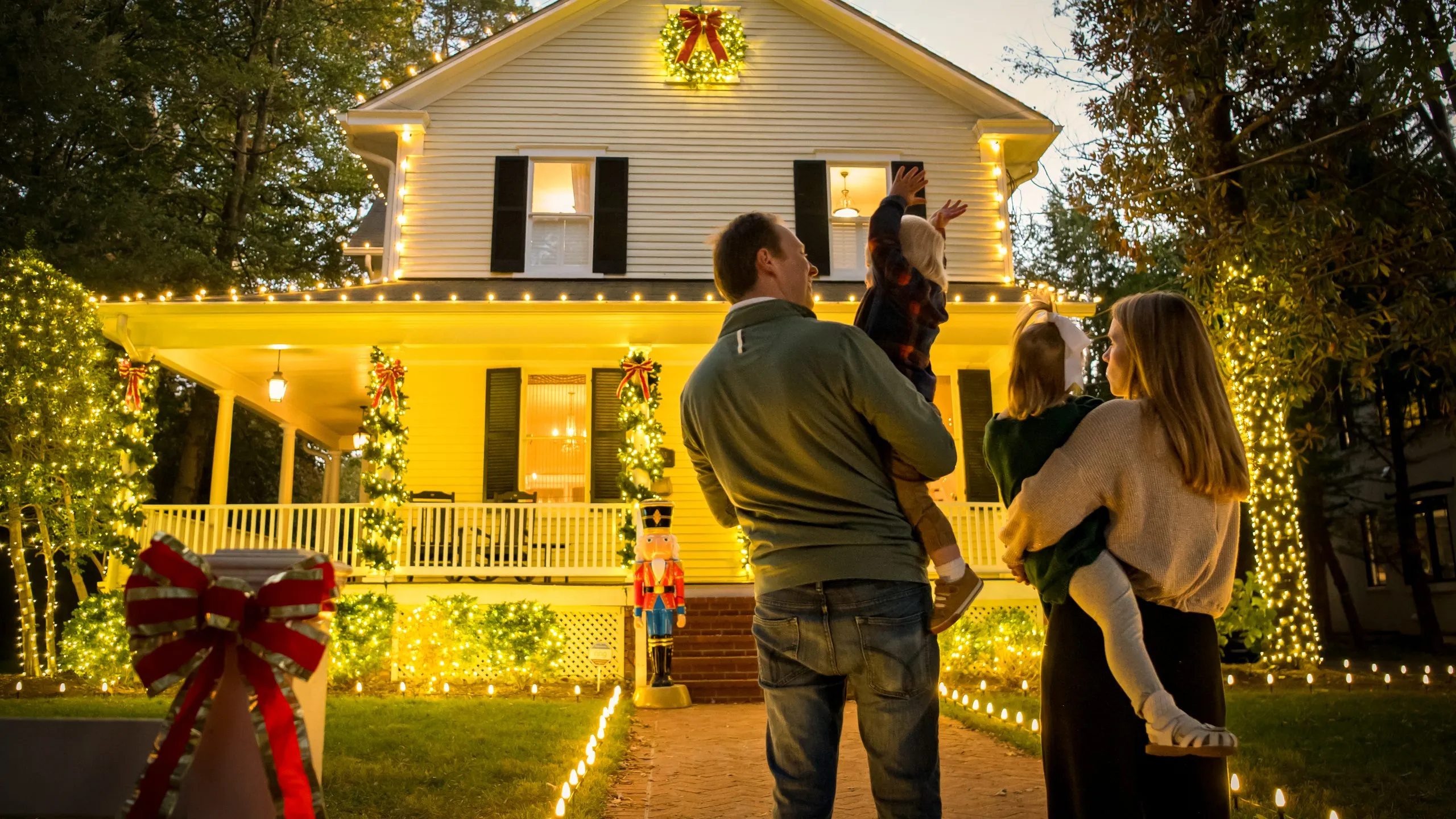 A family looking up at their lit two-story home, with garland, wreaths and a nutcracker beside the porch steps